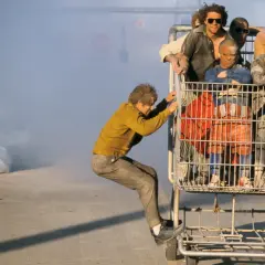 Grupa ludzi, jak prawdziwi jackass, pcha wózek sklepowy z pasażerami, w tle dym.