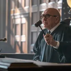 Starszy mężczyzna w todze sędziowskiej i okularach trzyma młotek sędziowski.