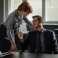 Kobieta z rudymi włosami podaje mężczyźnie w garniturze kartkę z rysunkiem. Atmosfera jak w filmie noir.