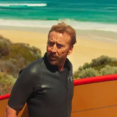 Nicolas Cage w piance z deską surfingową na plaży, jak w jednym z jego filmów.