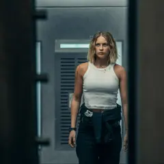 Kobieta w białej koszulce i ciemnych spodniach, z blizną na ramieniu, idzie korytarzem. Może to scena z jednego z topowych **Netflix filmy ranking**.