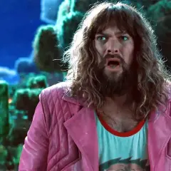 Jason Momoa w różowej marynarce i z długimi włosami, w tle pikselowe postacie. Jego filmowe i serialowe role są niezapomniane.