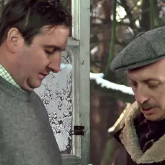Kargul i Pawlak rozmawiają przy zamarzniętym oknie. Jeden w szarym swetrze, drugi w czapce i kożuchu.