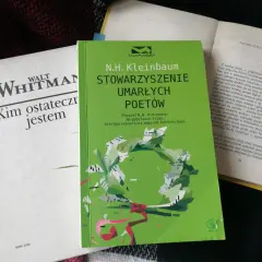 Zielona okładka książki "Stowarzyszenie Umarłych Poetów" N.H. Kleinbauma. Obok biała książka Walta Whitmana.