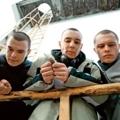 Trzech młodych mężczyzn, jakby z obsady "Jesteś Bogiem", pochyla się nad barierką. Jeden z nich trzyma w dłoniach jakiś przedmiot.