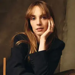 Maya Hawke w czarnej marynarce, opierająca głowę na dłoni. Aktorka znana z filmów, seriali i programów.