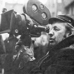 Mężczyzna w czapce i płaszczu patrzy przez obiektyw kamery filmowej.