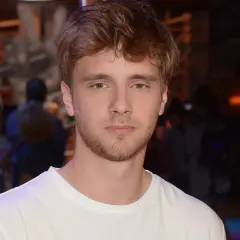 Młody mężczyzna z zarostem, w białej koszulce, przypomina aktora znanego z wielu maciej musiał filmy seriale i programy.