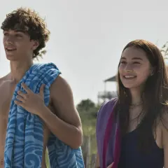 Uśmiechnięci nastolatkowie na plaży, jak z filmu "Tego lata stałam się piękna". Chłopak z kręconymi włosami i niebieskim ręcznikiem, dziewczyna z długimi brązowymi włosami i fioletowym ręcznikiem.