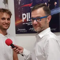 Mężczyzna w białej koszuli z niebieskimi okularami trzyma mikrofon, udziela wywiadu młodemu, uśmiechniętemu czarneckiemu aktorowi na tle plakatów teatralnych.