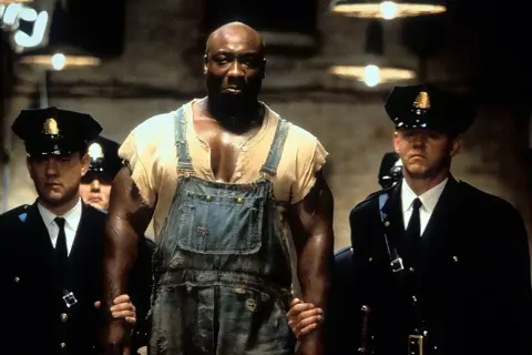 John Coffey w ogrodniczkach, prowadzony przez strażników więziennych. Scena z filmu "Zielona mila 2".