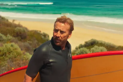 Nicolas Cage w piance z deską surfingową na plaży, jak w jednym z jego filmów.