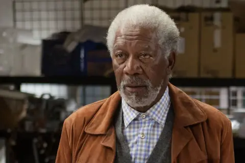 Morgan Freeman w brązowej kurtce i kratowanej koszuli, jak z jego filmów.