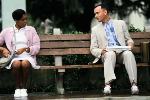 Forest Gump siedzi na ławce z walizką, obok niego siedzi kobieta.