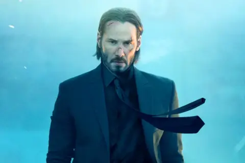 John Wick 5: Keanu Reeves w garniturze, z bliznami na twarzy, wpatruje się w obiektyw na tle błękitnego, mglistego nieba.