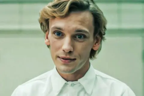 Jamie Campbell Bower, aktor wcielający się w postać Vecny, z niebieskimi oczami i blond włosami, w białej koszuli.