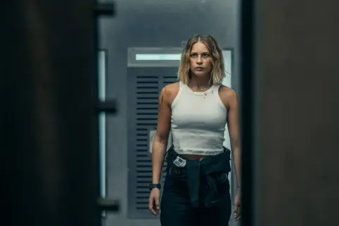 Kobieta w białej koszulce i ciemnych spodniach, z blizną na ramieniu, idzie korytarzem. Może to scena z jednego z topowych **Netflix filmy ranking**.