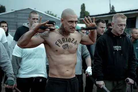 Groźny mężczyzna z tatuażem "Furioza" na klatce piersiowej, otoczony przez grupę mężczyzn. Czy to zapowiedź Furiozy 2, kiedy?