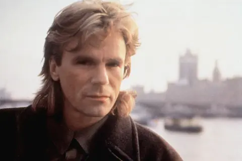 MacGyver obsada: Richard Dean Anderson jako MacGyver, z charakterystyczną fryzurą i w płaszczu, na tle mglistego miasta.