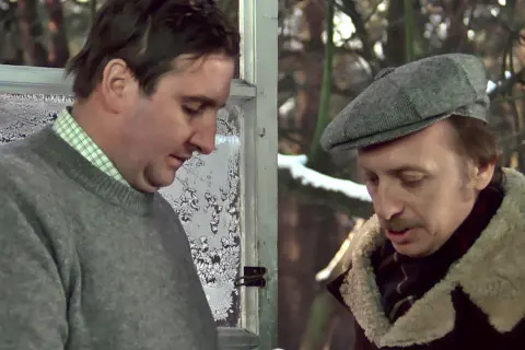 Kargul i Pawlak rozmawiają przy zamarzniętym oknie. Jeden w szarym swetrze, drugi w czapce i kożuchu.