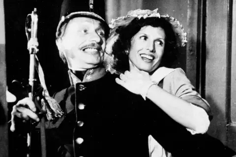 Uśmiechnięta para z filmu "C.K. Dezerterzy" - mężczyzna w mundurze i kobieta w kapeluszu.