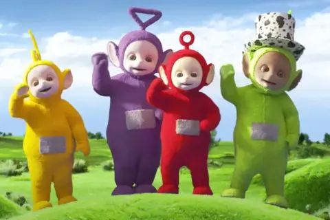 Tinky Winky, Dipsy, Laa-Laa i Po – teletubisie imiona znane dzieciom. Wesołe postacie machają do widzów na zielonej łące.