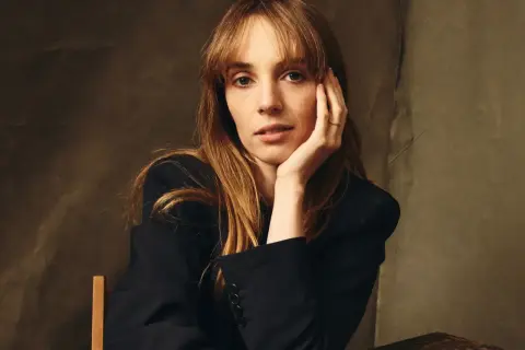 Maya Hawke w czarnej marynarce, opierająca głowę na dłoni. Aktorka znana z filmów, seriali i programów.