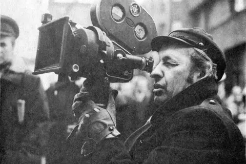 Mężczyzna w czapce i płaszczu patrzy przez obiektyw kamery filmowej.