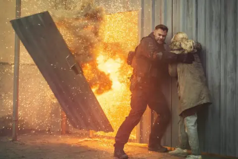 Mężczyzna w kamizelce taktycznej chroni kobietę przed eksplozją, jak w najlepszych filmach akcji.