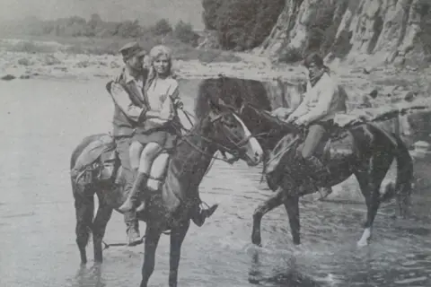 Para z "Wilczych ech" na koniach w rzece. W tle skaliste zbocze i drzewa.