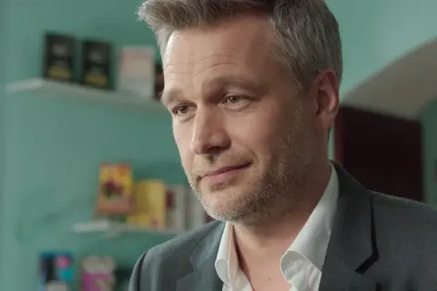 Michał Żebrowski w jednym z filmów, z siwiejącymi włosami i zarostem, w białej koszuli i szarym garniturze.