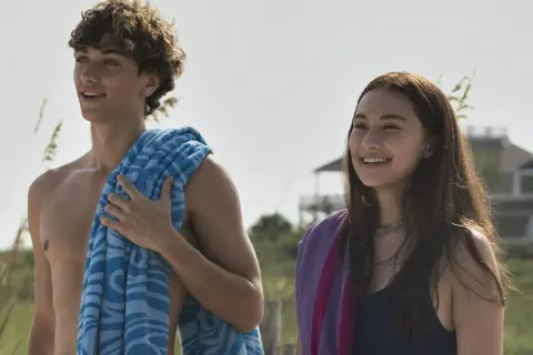 Uśmiechnięci nastolatkowie na plaży, jak z filmu "Tego lata stałam się piękna". Chłopak z kręconymi włosami i niebieskim ręcznikiem, dziewczyna z długimi brązowymi włosami i fioletowym ręcznikiem.