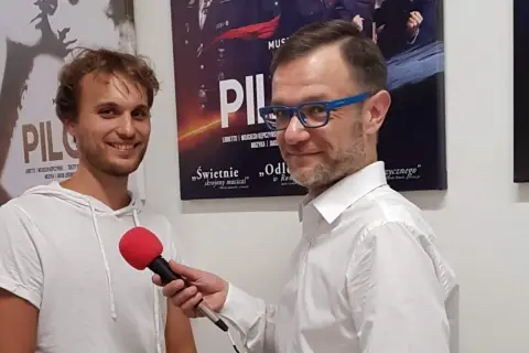Mężczyzna w białej koszuli z niebieskimi okularami trzyma mikrofon, udziela wywiadu młodemu, uśmiechniętemu czarneckiemu aktorowi na tle plakatów teatralnych.