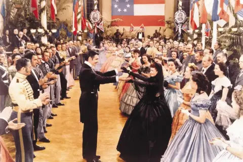 Scarlett O'Hara i Rhett Butler tańczą na balu. W tle widać całą obsadę filmu "Przeminęło z wiatrem" w historycznych strojach.