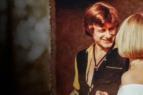 Uśmiechnięty mężczyzna w stylu lat 70. rozmawia z kobietą. Scena jak z filmu "Tulipan".