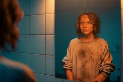 Mroczna scena z filmu "Morderczynie" - młoda kobieta w poplamionej bluzie patrzy w lustro, jej twarz wyraża niepokój.