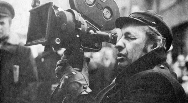 Mężczyzna w czapce i płaszczu patrzy przez obiektyw kamery filmowej.