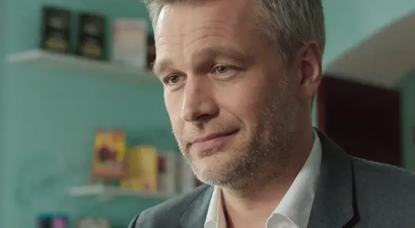 Michał Żebrowski w jednym z filmów, z siwiejącymi włosami i zarostem, w białej koszuli i szarym garniturze.
