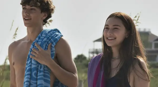 Uśmiechnięci nastolatkowie na plaży, jak z filmu "Tego lata stałam się piękna". Chłopak z kręconymi włosami i niebieskim ręcznikiem, dziewczyna z długimi brązowymi włosami i fioletowym ręcznikiem.