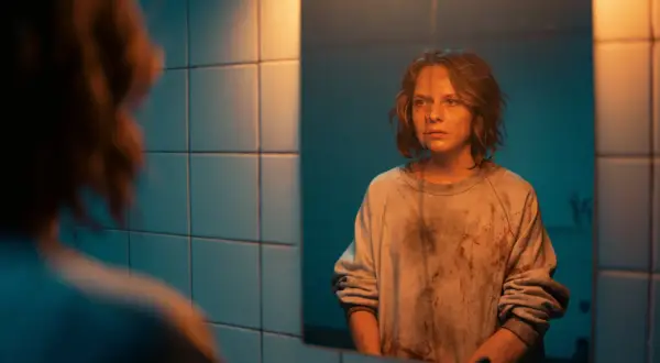 Mroczna scena z filmu "Morderczynie" - młoda kobieta w poplamionej bluzie patrzy w lustro, jej twarz wyraża niepokój.
