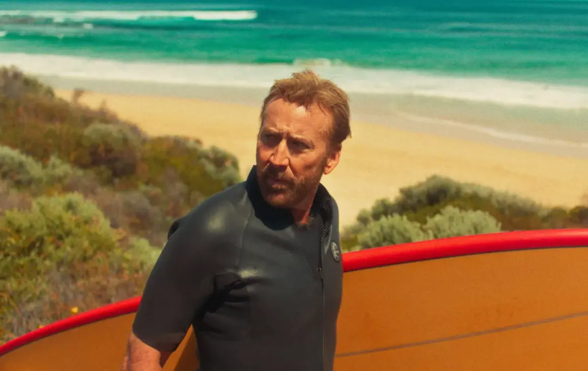 Nicolas Cage w piance z deską surfingową na plaży, jak w jednym z jego filmów.
