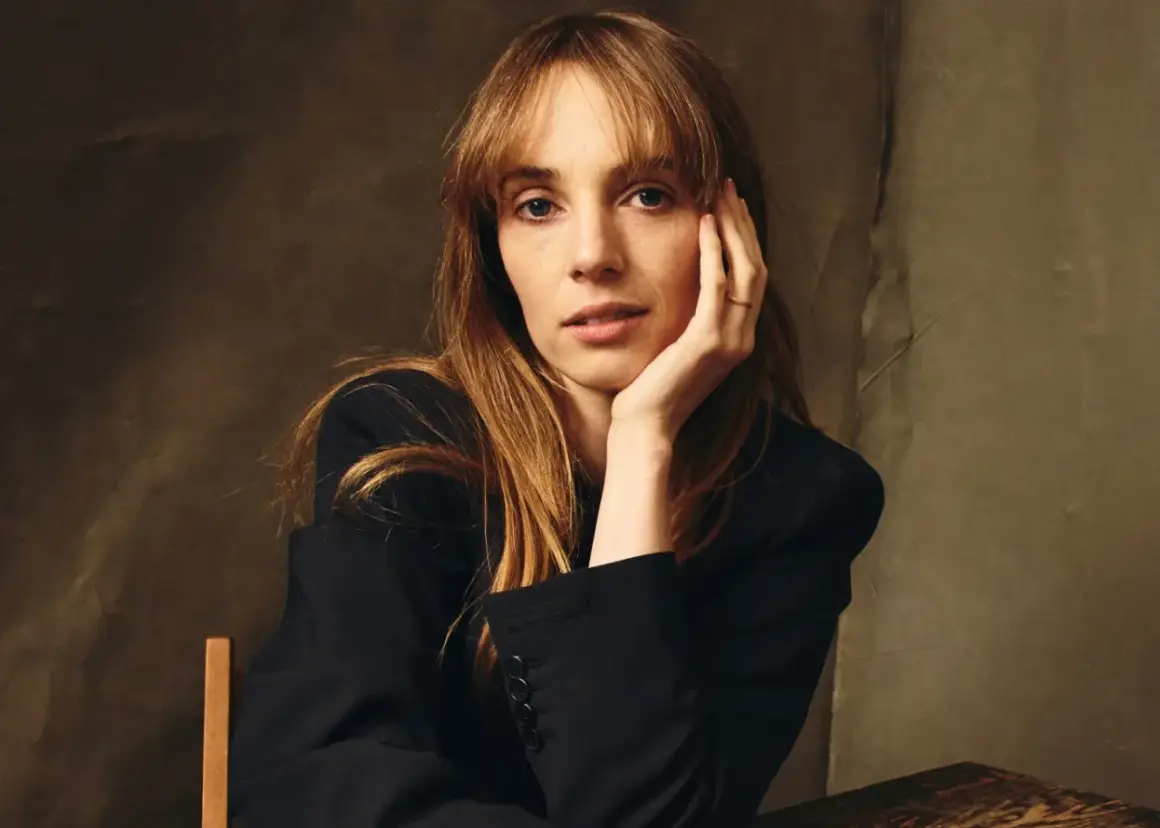 Maya Hawke w czarnej marynarce, opierająca głowę na dłoni. Aktorka znana z filmów, seriali i programów.