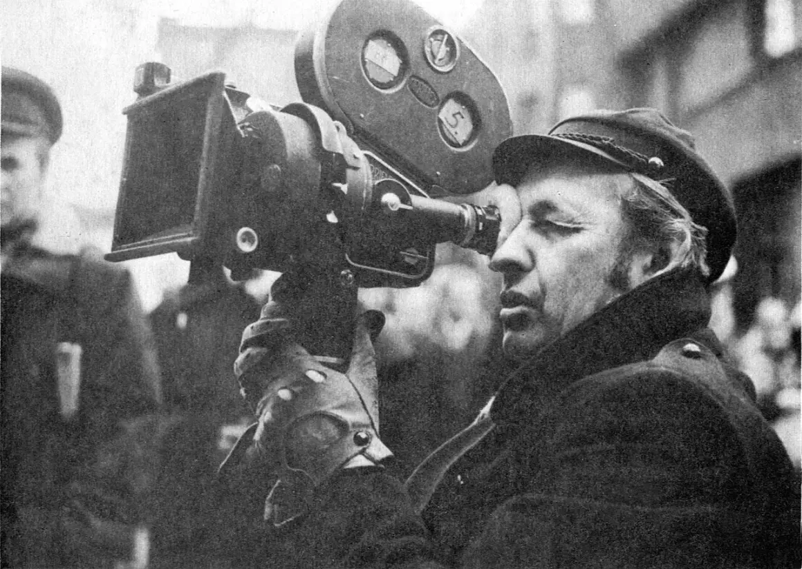 Mężczyzna w czapce i płaszczu patrzy przez obiektyw kamery filmowej.