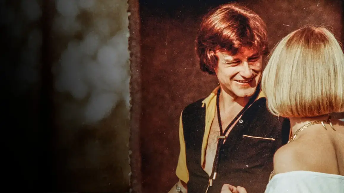 Uśmiechnięty mężczyzna w stylu lat 70. rozmawia z kobietą. Scena jak z filmu "Tulipan".