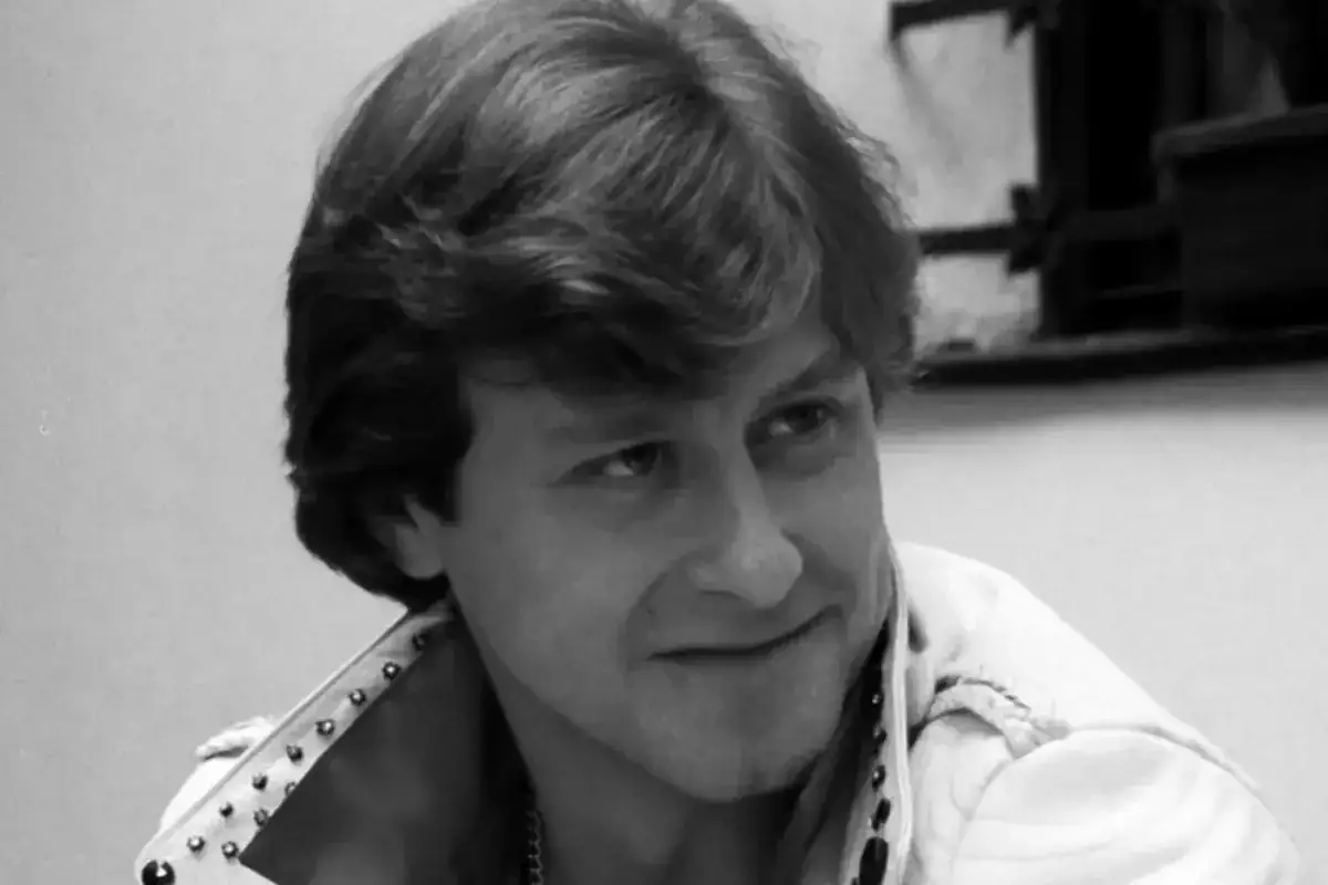 Mężczyzna z burzą włosów, w białej kurtce z ćwiekami, uśmiecha się lekko. Kadr z filmu 