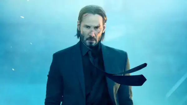 John Wick 5: Keanu Reeves w garniturze, z bliznami na twarzy, wpatruje się w obiektyw na tle błękitnego, mglistego nieba.