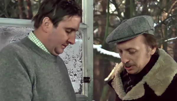 Kargul i Pawlak rozmawiają przy zamarzniętym oknie. Jeden w szarym swetrze, drugi w czapce i kożuchu.