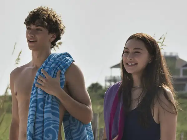 Uśmiechnięci nastolatkowie na plaży, jak z filmu "Tego lata stałam się piękna". Chłopak z kręconymi włosami i niebieskim ręcznikiem, dziewczyna z długimi brązowymi włosami i fioletowym ręcznikiem.