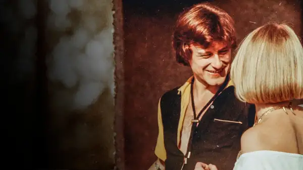 Uśmiechnięty mężczyzna w stylu lat 70. rozmawia z kobietą. Scena jak z filmu "Tulipan".